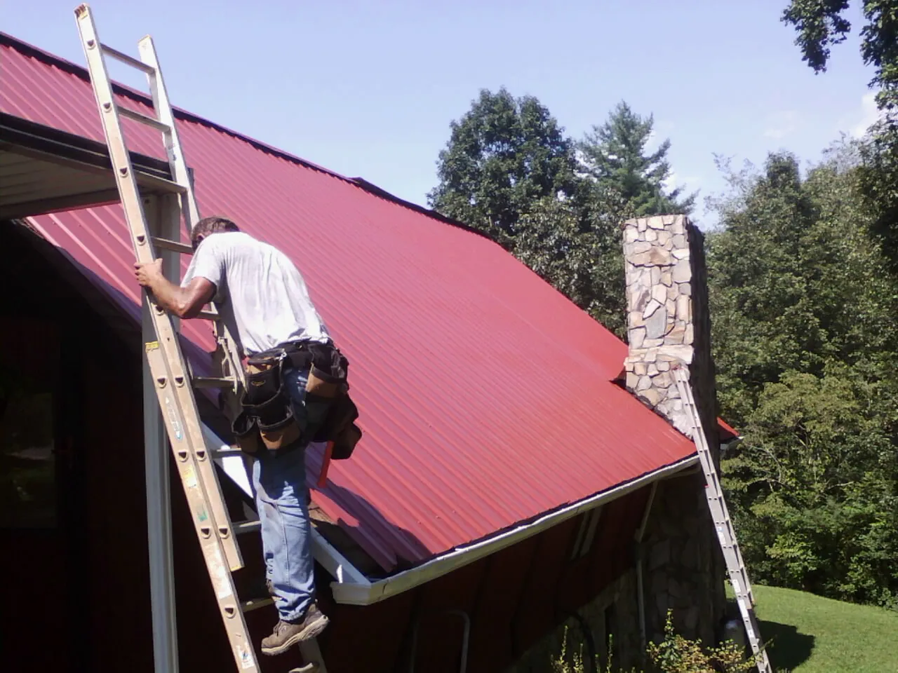 Roofer on a ladder beside a red metal roof and stone chimney — Soffit & Fascia Repair by Roofing Brothers in Earlsboro